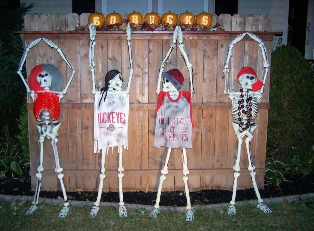 Four skeletons dressed in Ohio State Buckeyes shirts and hats with arms raised along a wooden fence, below pumpkins spelling GO BUCKS