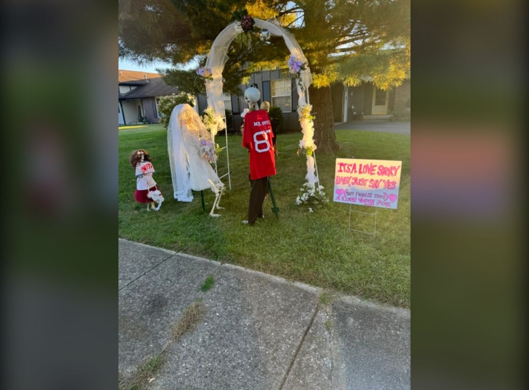 Skeleton wedding under a flowered arch: a skeleton bride in a veil, a groom in a red number 8 jersey and a flower girl, beside a sign reading It’s a Love Story, Baby Just Say Yes