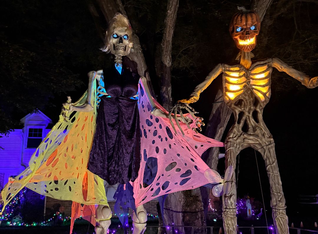 Night display: a skeleton in a purple dress with glowing pink and yellow wings beside a giant pumpkin-headed skeleton with a glowing ribcage, with a sign reading MY MAN, surrounded by purple lights