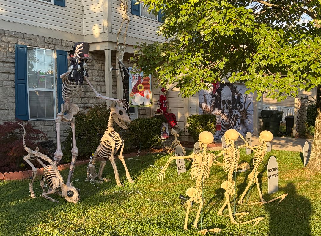 Top-hatted skeleton with a scythe walking a skeleton dog and cat across a front lawn, with skeletons crawling toward a row of tombstones